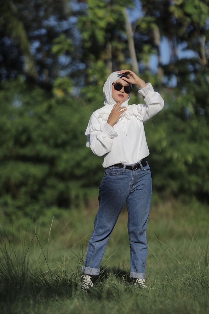 Fashionable woman in jeans and white blouse posing outdoors with sunglasses and headscarf.