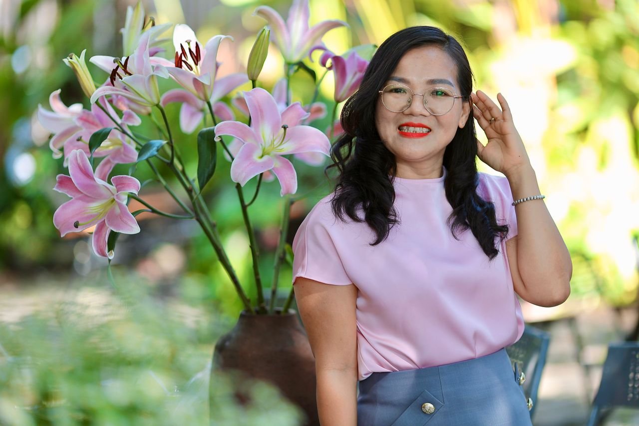 Smiling woman in garden with pink lilies, wearing glasses and pink blouse.