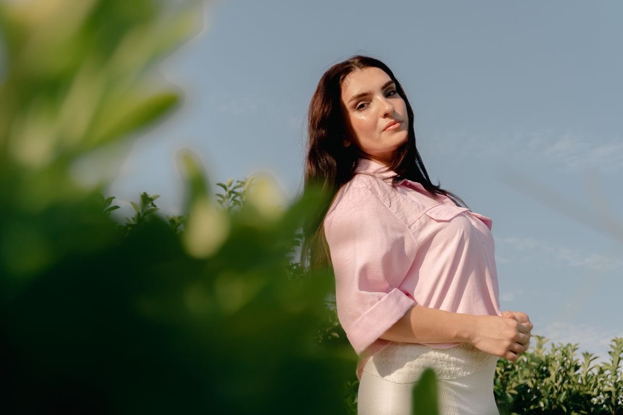 A serene portrait of a woman in a pink blouse standing amidst greenery, basking in natural sunlight.