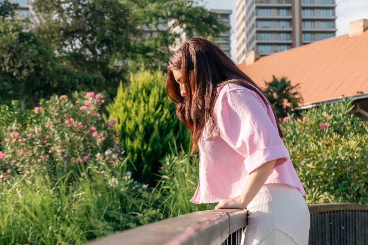 A woman in a pink blouse gazes over a balcony at a vibrant garden with blooming flowers.